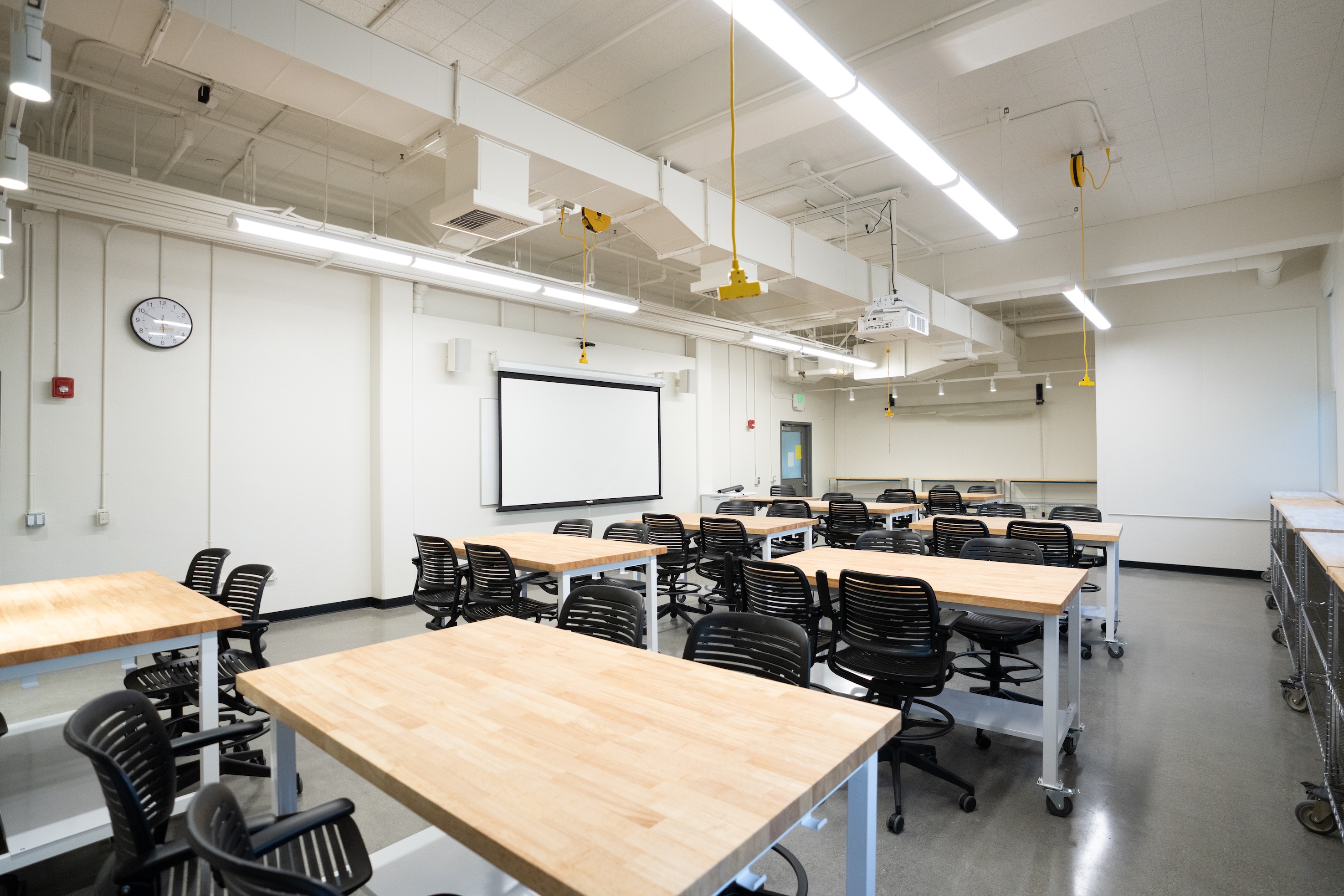 Bright modern classroom with wooden worktables, multiple black rolling chairs and whiteboard.