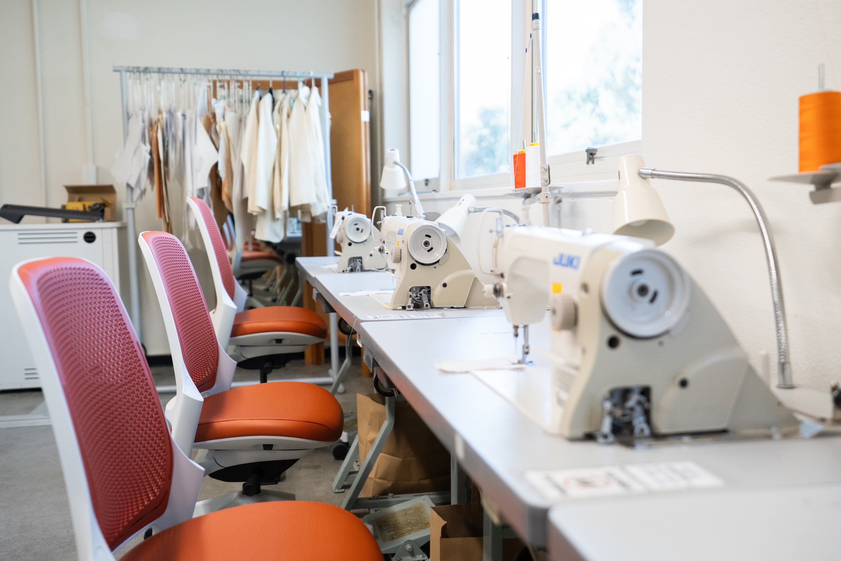 Row of white sewing machines and orange-red chairs in a bright sewing studio.
