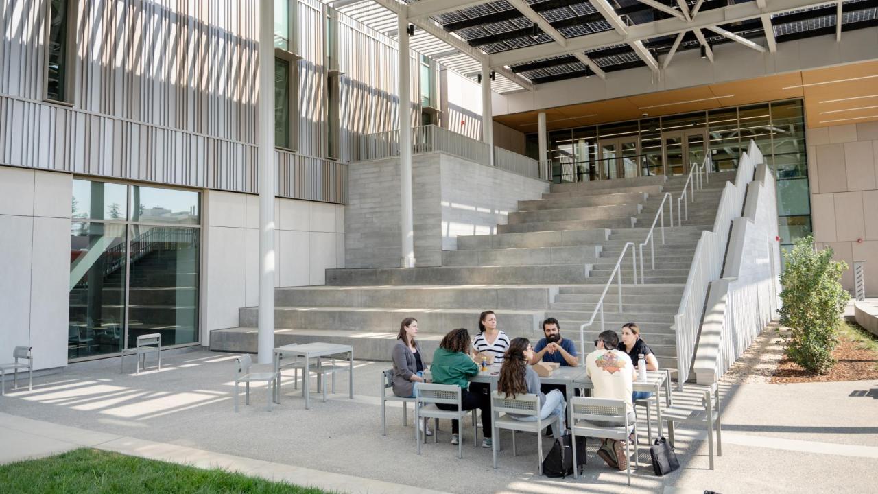 Group of people sit at a table near the large staircase outside the Teaching and Learning Complex