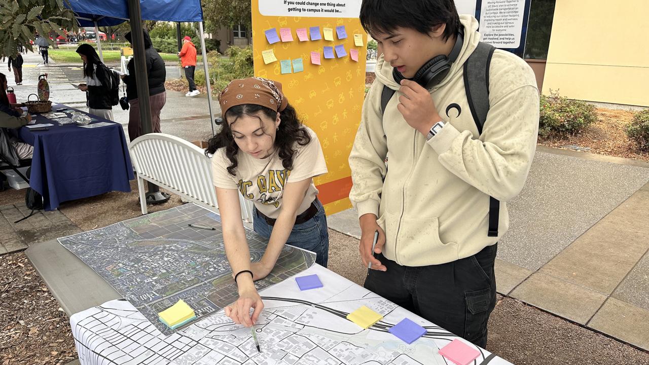 A young woman points to a place on a map while a young man looks on. They are at an outdoor tabling event about transporatation planning.