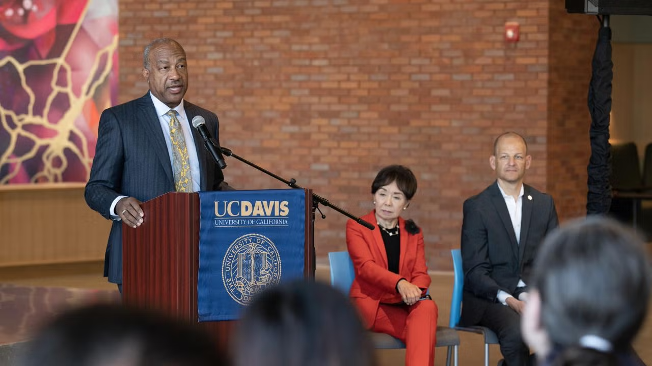 Standing at a lectern, UC Davis Chancellor Gary S. May speaks into a microphone as a seated man and woman look on